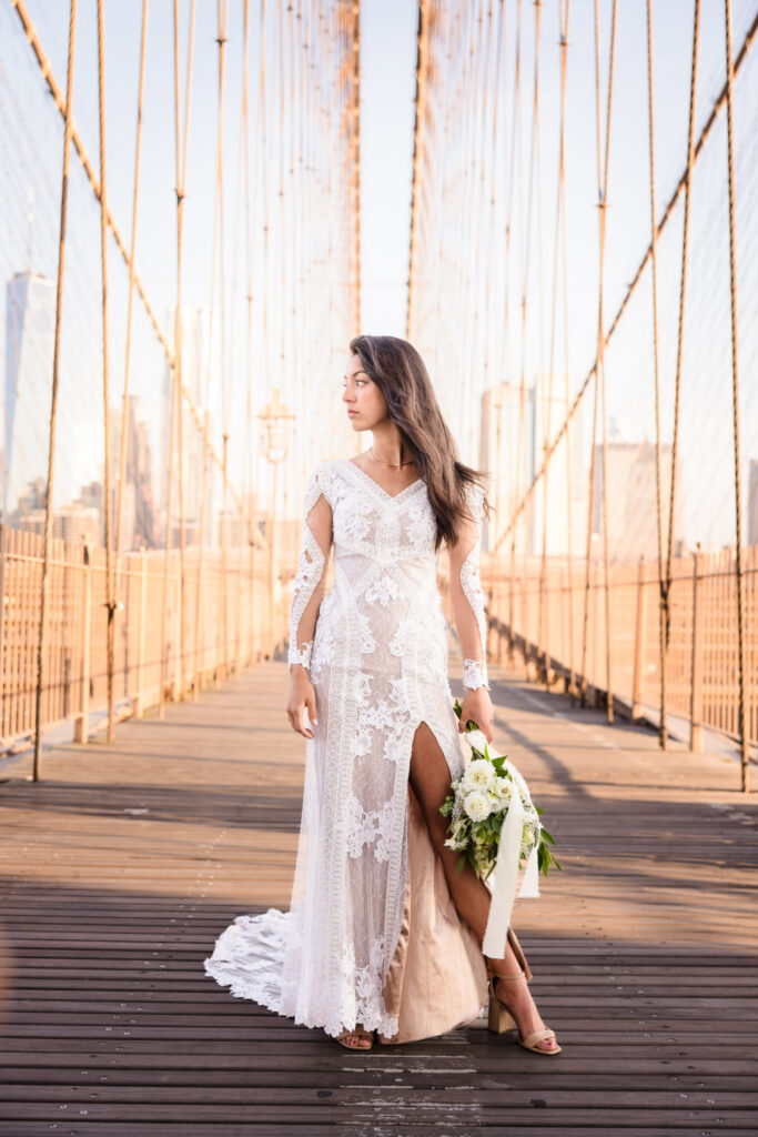 Elegant sunrise bridal portrait on the Brooklyn Bridge, bride standing centered between suspension cables holding a white and green bouquet, her lace gown glowing in warm July morning light.