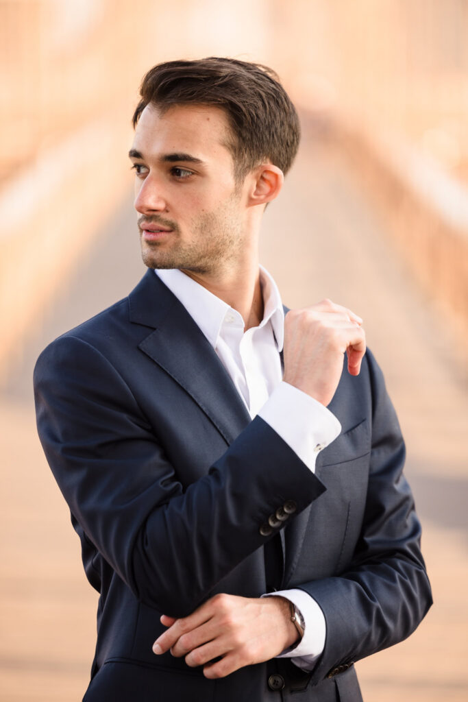 Close-up sunrise groom portrait, adjusting his navy suit jacket with soft golden July morning light and suspension cables blurred in the background.
