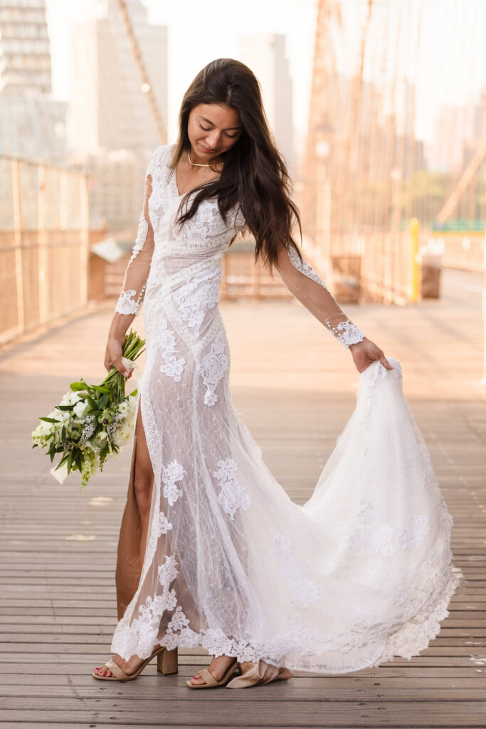 Sunrise Brooklyn Bridge bridal portrait of bride looking down while holding her flowing lace train and bouquet, her gown glowing in soft golden July morning light along the wooden walkway.