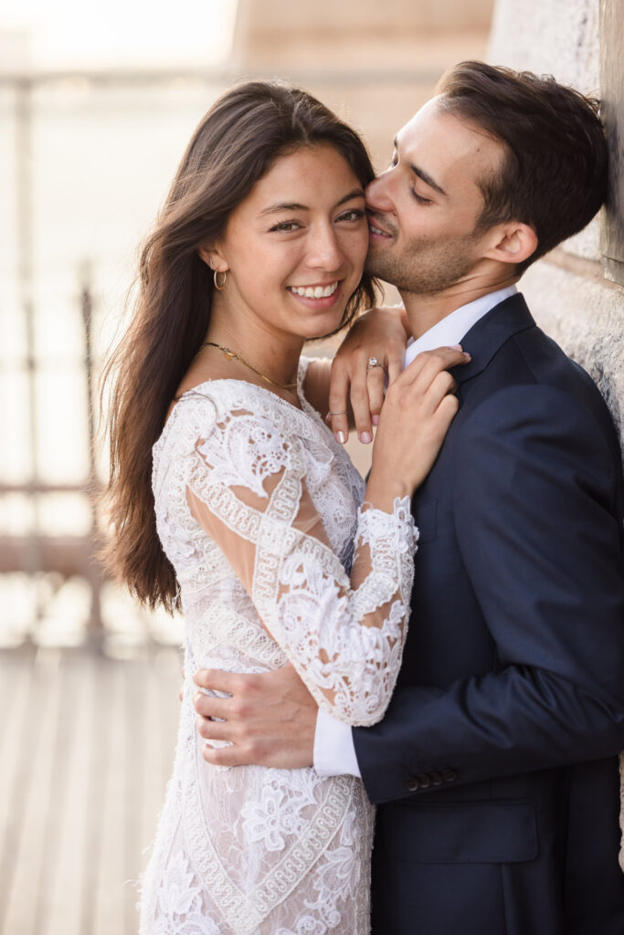 Romantic close-up of couple embracing against the Brooklyn Bridge stone arches, featuring a bride in a lace dress and groom in a navy suit for elegant NYC engagement photos.