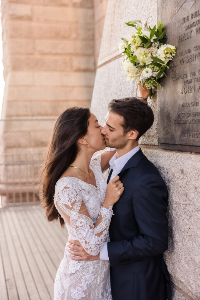 Romantic wedding portrait of bride and groom kissing against the stone pillar beneath the Roebling plaque, bouquet lifted overhead and warm July morning light illuminating the scene.