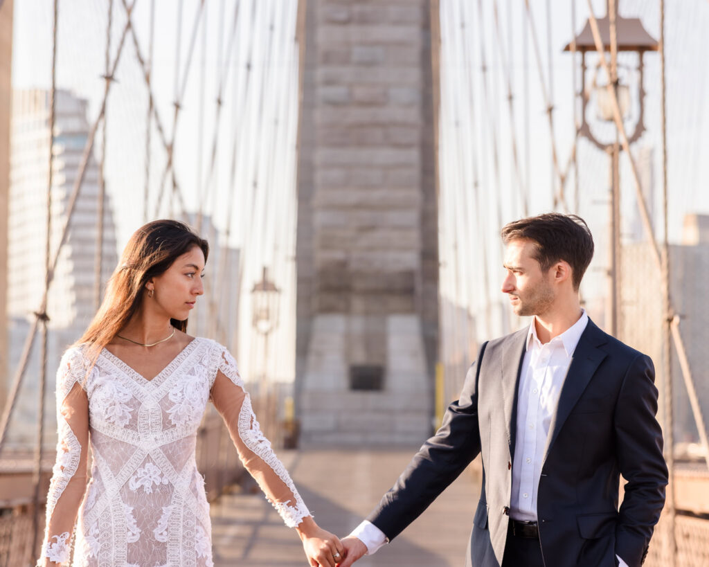 Elegant couple holding hands on the Brooklyn Bridge at golden hour, framed by the iconic stone arches and suspension cables for timeless NYC engagement photos.