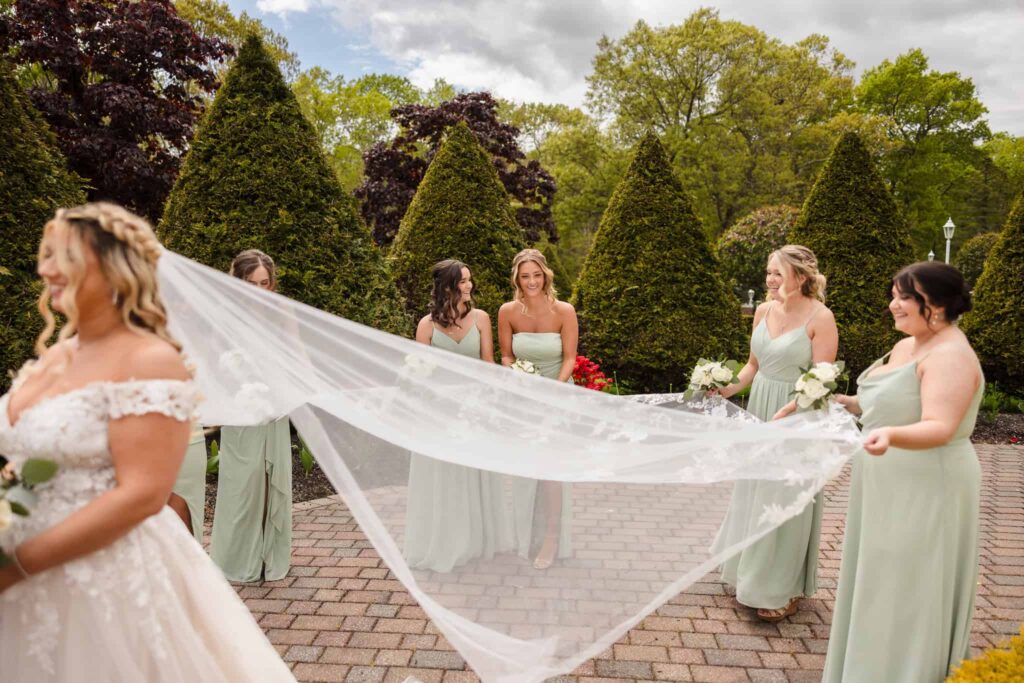 Bridesmaids in sage green dresses holding the bride’s flowing veil during a spring Vineyard at East Wind wedding, surrounded by manicured evergreens and garden pathways.