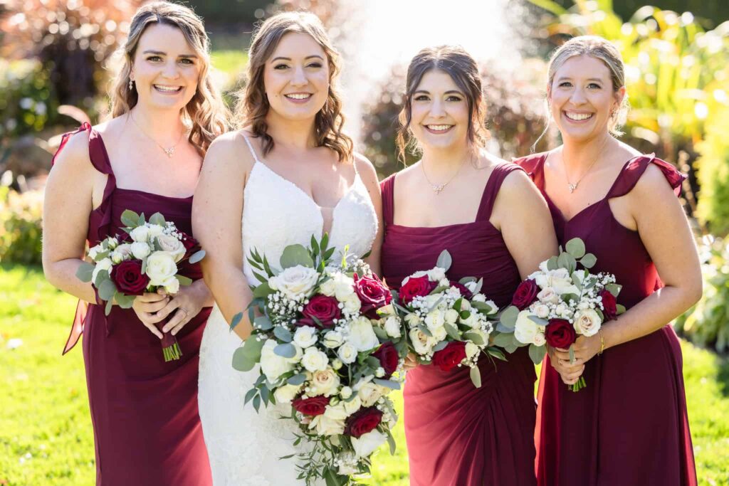 Bride and bridesmaids in burgundy dresses holding red and white rose bouquets during a fall Vineyard at East Wind wedding in Suffolk County, Long Island.