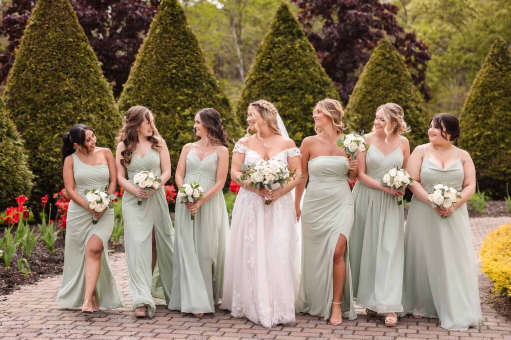 Bride walking with bridesmaids in sage green dresses along a garden path at the Vineyard at East Wind wedding venue, surrounded by spring florals and evergreens.