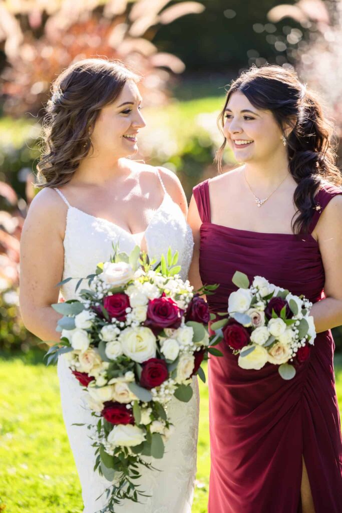 Bride and bridesmaid smiling at each other while holding red and white rose bouquets during a wedding on a sunny Long Island afternoon.
