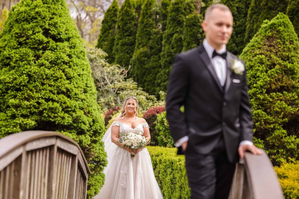 Bride approaching the groom for their first look across a wooden bridge surrounded by lush greenery at the Vineyard at East Wind wedding venue in Long Island.