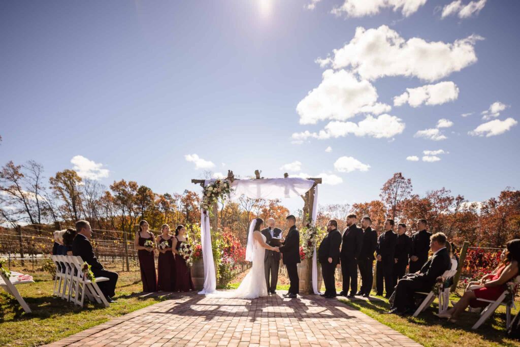 Outdoor fall ceremony at the Vineyard at East Wind wedding venue, with the couple exchanging vows beneath a floral arbor surrounded by vineyard views and clear blue skies.