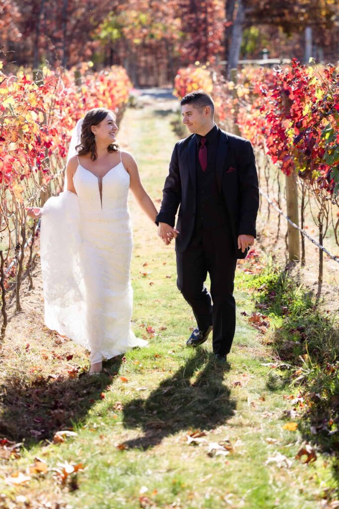 Bride and groom walking hand in hand through vibrant fall foliage at their Vineyard at East Wind wedding, showcasing Long Island vineyard scenery in autumn.
