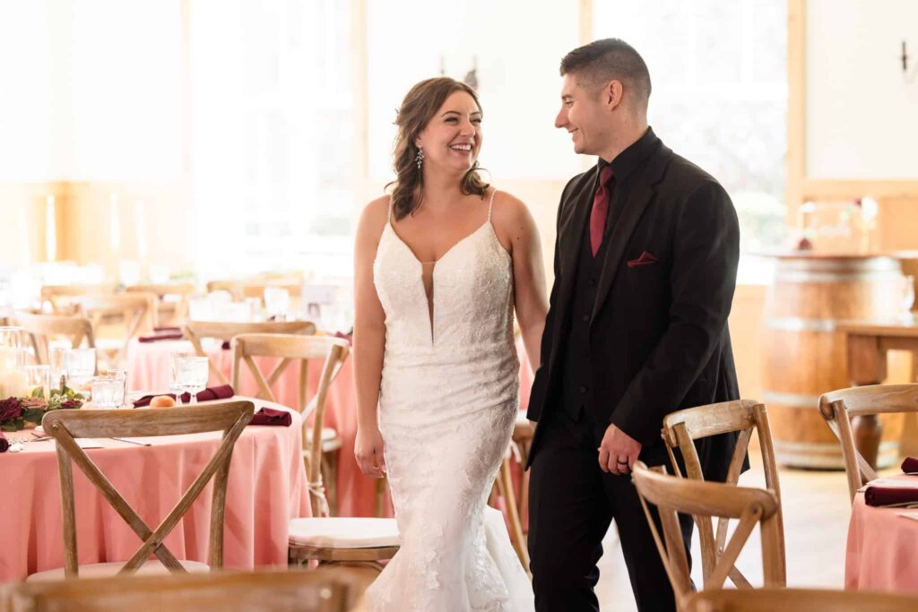 Bride and groom share a joyful moment walking through the reception space at their Vineyard at East Wind wedding, surrounded by rustic wood chairs and blush table settings.