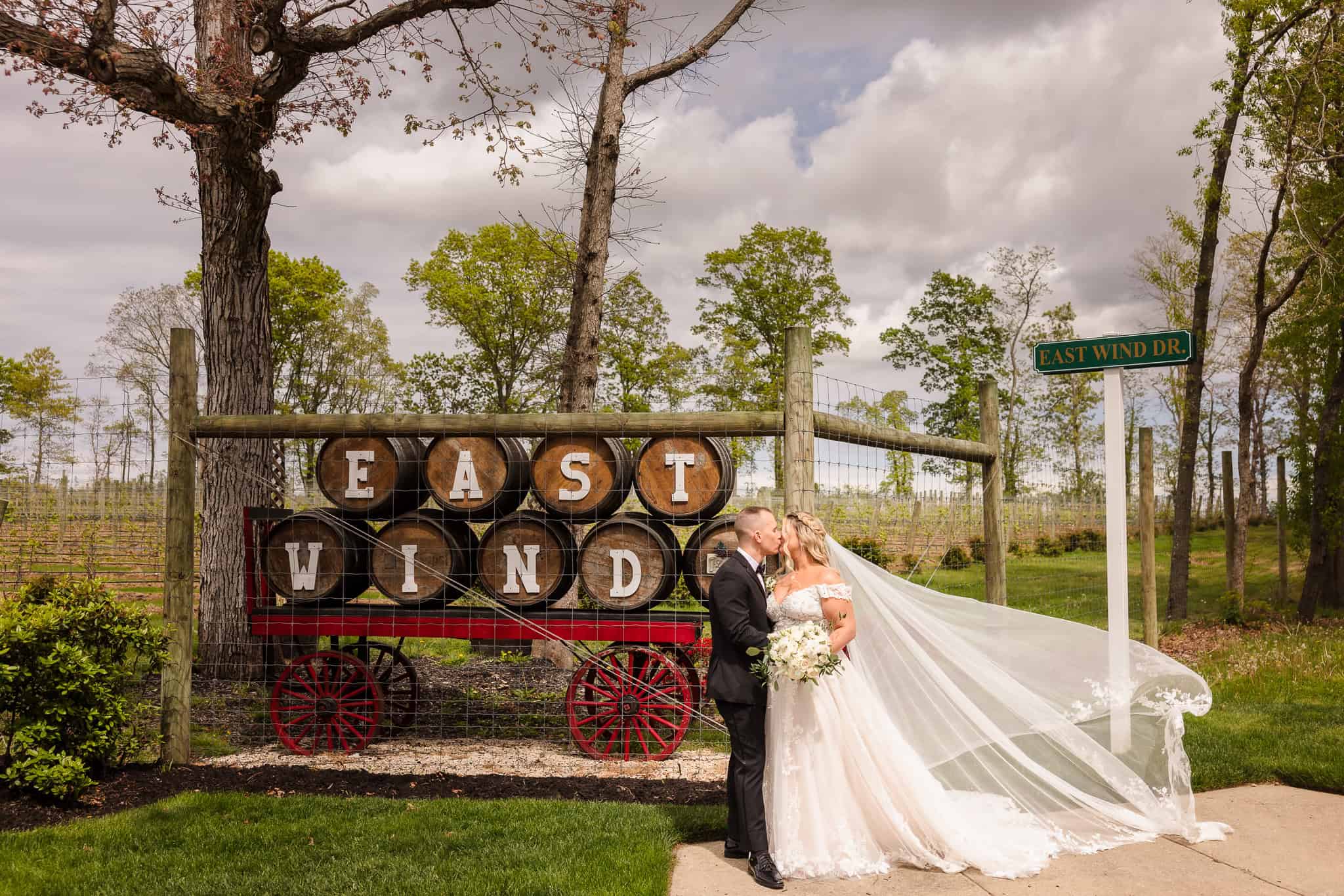 Bride and groom share a kiss in front of wine barrels spelling "East Wind" at their Vineyard at East Wind wedding, highlighting the scenic Long Island vineyard entrance.