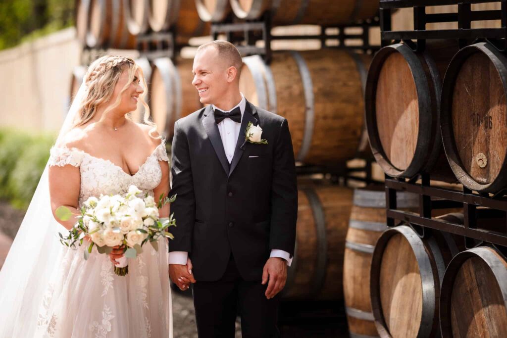 Bride and groom holding hands and smiling in front of stacked wine barrels at the Vineyard at East Wind wedding venue on Long Island.