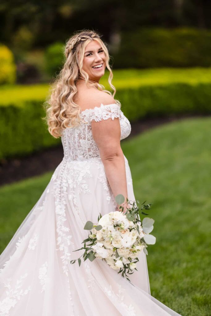 Bride smiling over her shoulder while holding a white rose bouquet, captured in the lush gardens of the Vineyard.