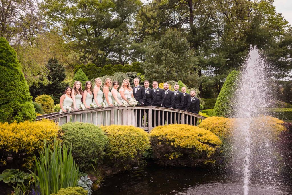 Bridal party posing on a wooden bridge over a garden pond with a fountain at the Vineyard at East Wind wedding venue.