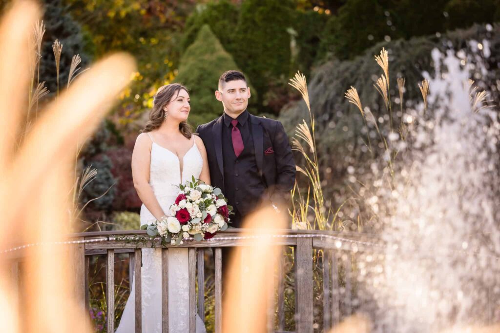 Bride and groom standing on a wooden bridge near a fountain, surrounded by ornamental grasses at the Vineyard at East Wind.