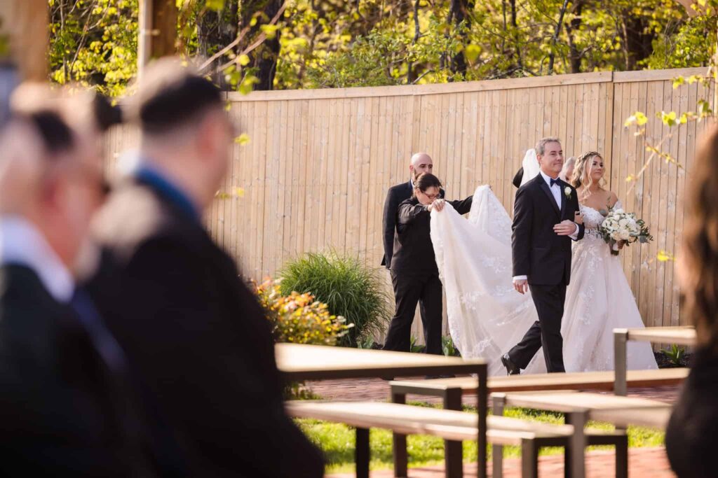 Bride walking down the aisle with her father at the Vineyard at East Wind, as attendants adjust her gown and guests look on.