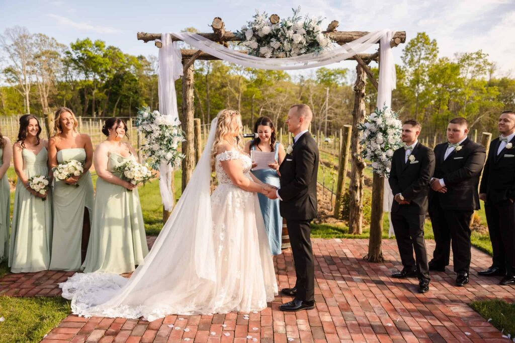 Bride and groom exchange vows under a rustic floral arch during their outdoor vineyard ceremony at East Wind, surrounded by their bridal party.