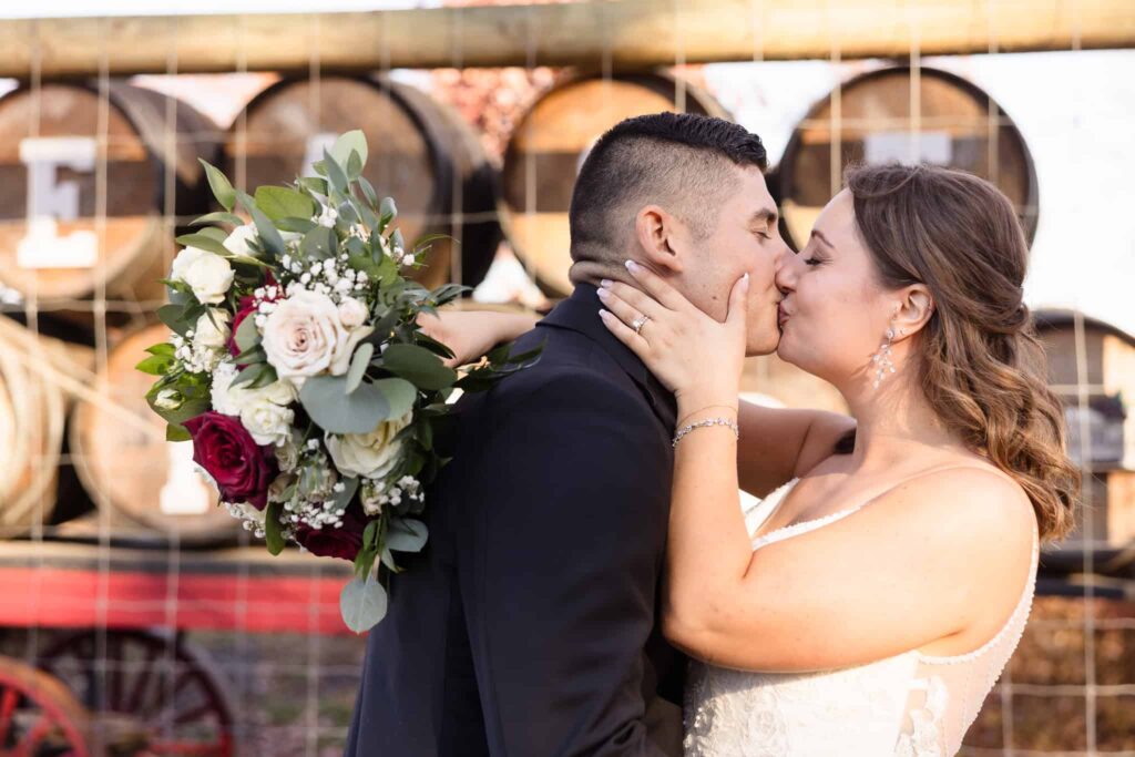 Bride and groom share a romantic kiss in front of rustic wine barrels at East Wind, with the bride holding a bouquet of red and white roses.