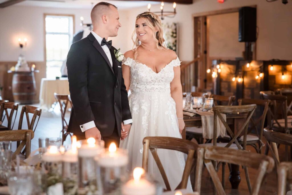 Bride and groom smile at each other while holding hands inside a warmly lit wedding reception venue with wooden chairs and floating candles.
