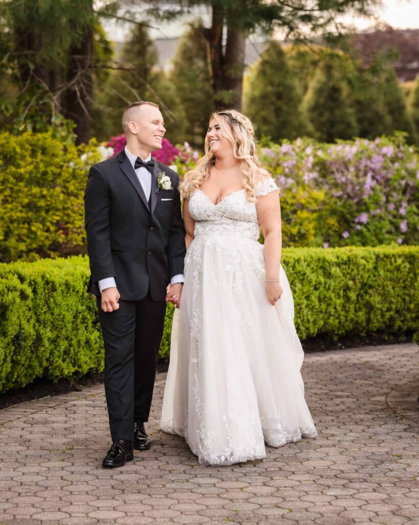 Bride and groom smiling at each other while walking hand-in-hand through a blooming garden path at the Vineyard at East Wind.
