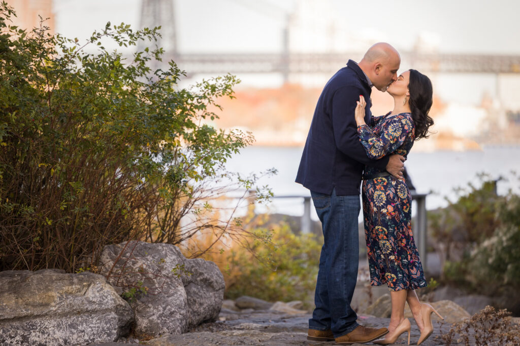 Winter engagement photos in DUMBO featuring Regina and Dominick sharing a kiss along the Brooklyn waterfront, framed by soft Brooklyn winter light and a subtle Manhattan Bridge backdrop during their off-season engagement session NYC.