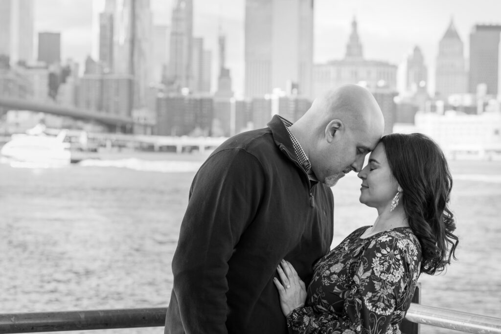 Black and white portrait of couple embracing along the Brooklyn waterfront with skyline backdrop during a quiet early morning engagement session in DUMBO.