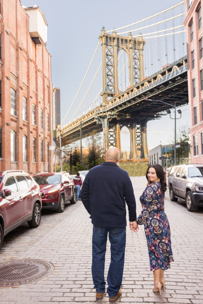 Winter engagement photos in DUMBO featuring Regina and Dominick walking hand in hand along the cobblestone streets DUMBO is known for, framed by Washington Street Manhattan Bridge views and classic brick and iron textures.