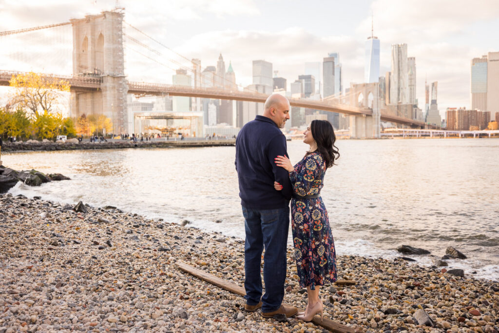 Winter engagement photos in DUMBO featuring Regina and Dominick standing along the rocky Brooklyn waterfront, with the Brooklyn Bridge and Lower Manhattan skyline creating a clean architectural engagement photos Brooklyn setting in soft winter light.