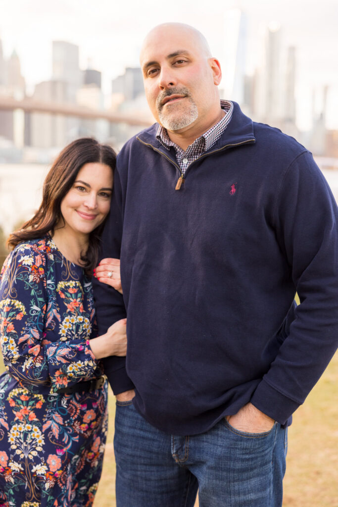 Couple embracing at Brooklyn Bridge Park with the Manhattan skyline softly blurred behind them, highlighting a relaxed Brooklyn waterfront engagement session near DUMBO.