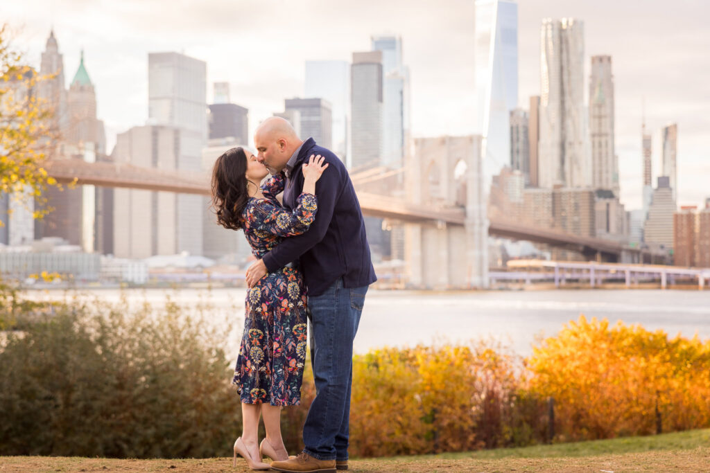 Couple embracing at Brooklyn Bridge Park during golden hour in their DUMBO engagement session, with the Brooklyn Bridge and Lower Manhattan skyline creating warm, cinematic engagement photos in DUMBO Brooklyn.