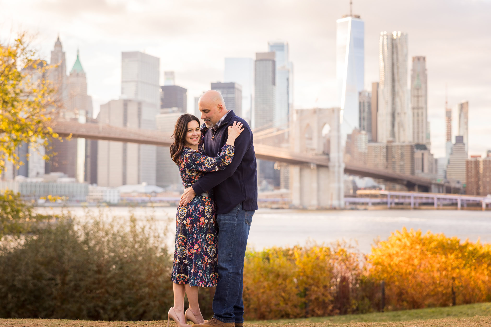 Winter engagement photos in DUMBO capturing Regina and Dominick embracing with the Brooklyn Bridge and Lower Manhattan skyline behind them, highlighting clean composition and soft Brooklyn winter light during their off-season engagement session NYC.