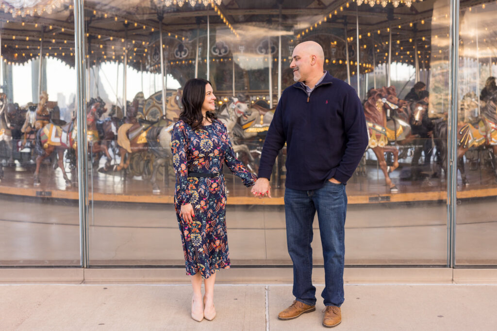 DUMBO engagement session featuring Regina and Dominick holding hands in front of Jane’s Carousel, blending architectural engagement photos Brooklyn couples love with clean winter tones and refined urban engagement photos NYC style.