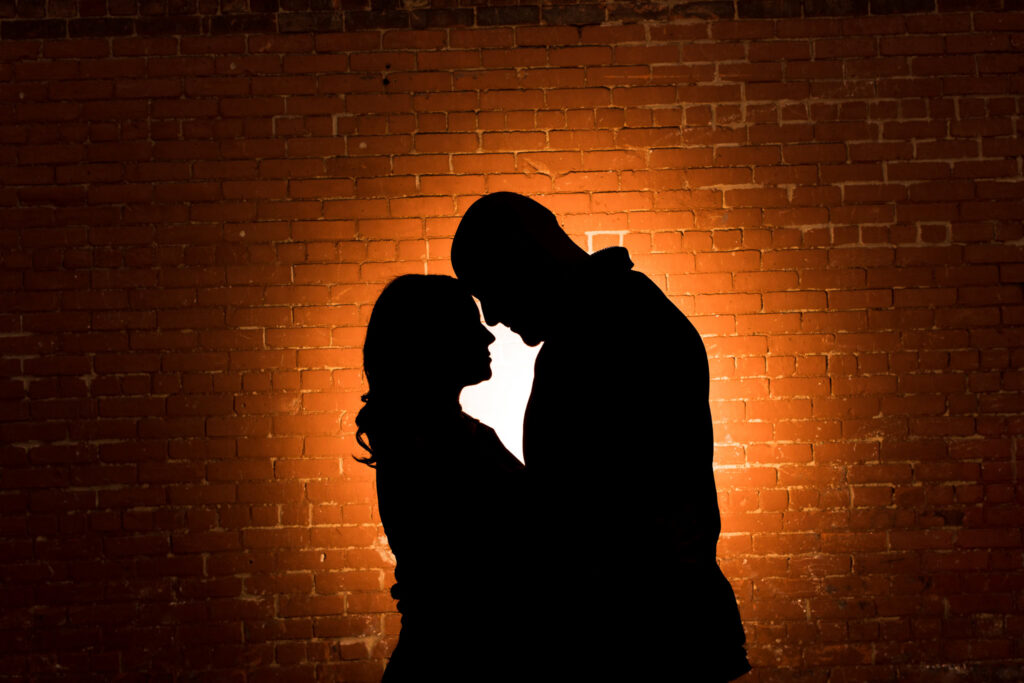 Engagement photos featuring Regina and Dominick in a dramatic silhouette against a glowing brick wall, highlighting rich brick textures and refined urban engagement photos NYC composition.