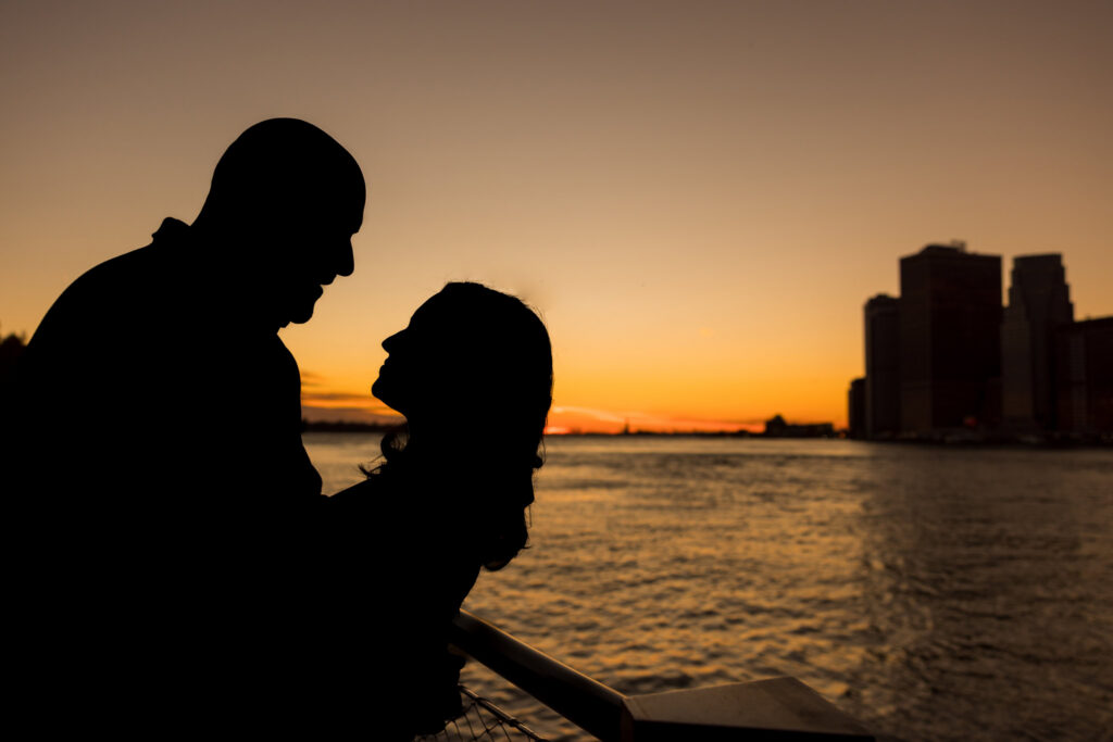 Winter engagement photos in DUMBO capturing Regina and Dominick in silhouette at sunset along the Brooklyn waterfront, with warm sky tones and the Lower Manhattan skyline creating refined urban engagement photos NYC couples love.