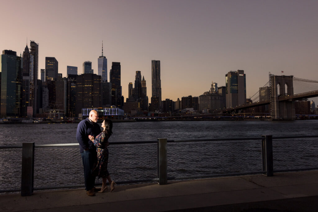 Winter engagement photos in DUMBO capturing Regina and Dominick at twilight along the Brooklyn waterfront, with the Lower Manhattan skyline and Brooklyn Bridge creating a dramatic architectural engagement photos Brooklyn backdrop.