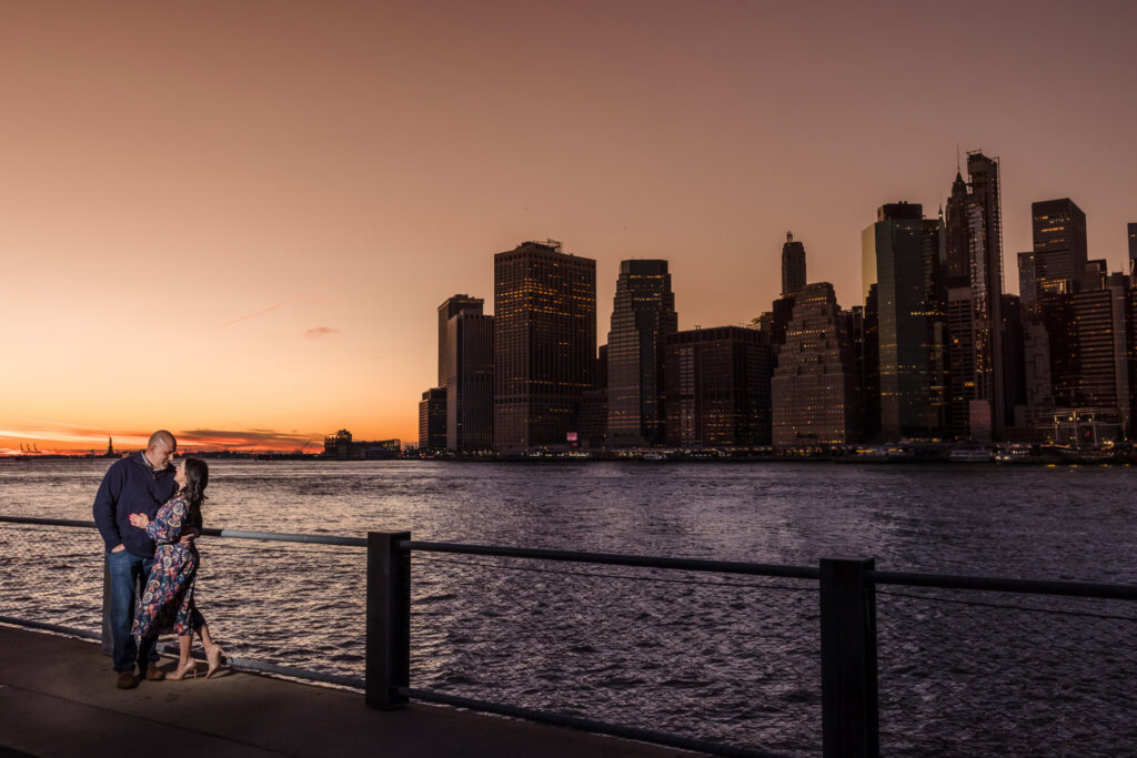 Winter engagement photos in DUMBO capturing Regina and Dominick at sunset along the Brooklyn waterfront, with sweeping Lower Manhattan skyline views and golden winter light defining refined urban engagement photos NYC couples cherish.