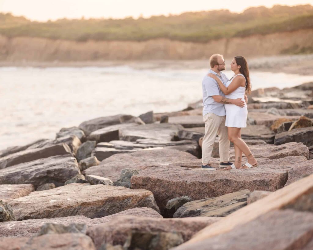 Couple embracing on the rocky shoreline during a Montauk engagement session, with soft golden hour light and coastal cliffs at Camp Hero State Park in the background.