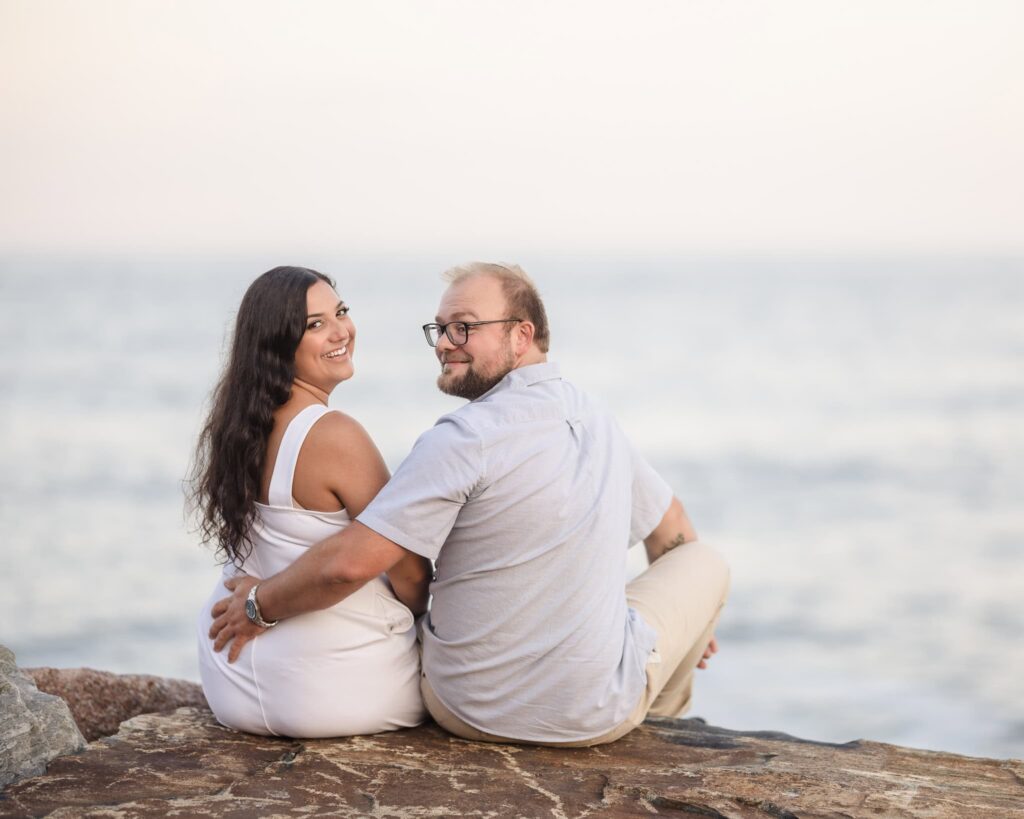 Engaged couple seated on coastal rocks during a Montauk engagement session, looking back toward the camera with soft ocean tones and evening light along the East End shoreline.