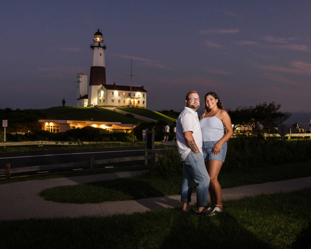 Couple posing at blue hour during a Montauk engagement session, with the illuminated Montauk Point Lighthouse glowing against the evening sky in the Hamptons.