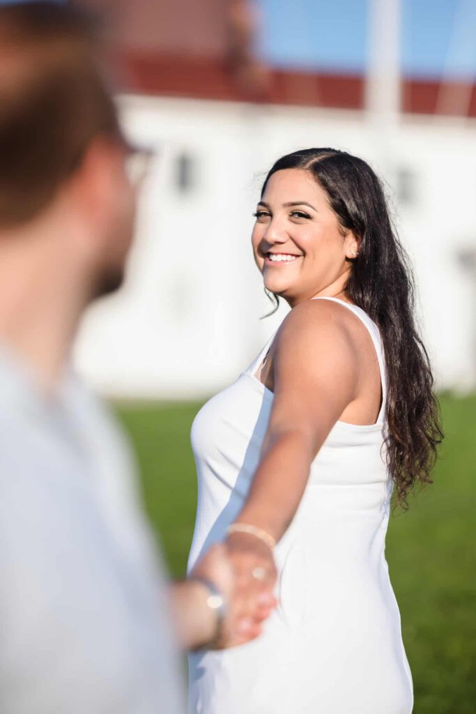 Bride-to-be smiling over her shoulder while leading her fiancé by the hand during a Montauk Point engagement session, with soft evening light and lighthouse grounds in the background.