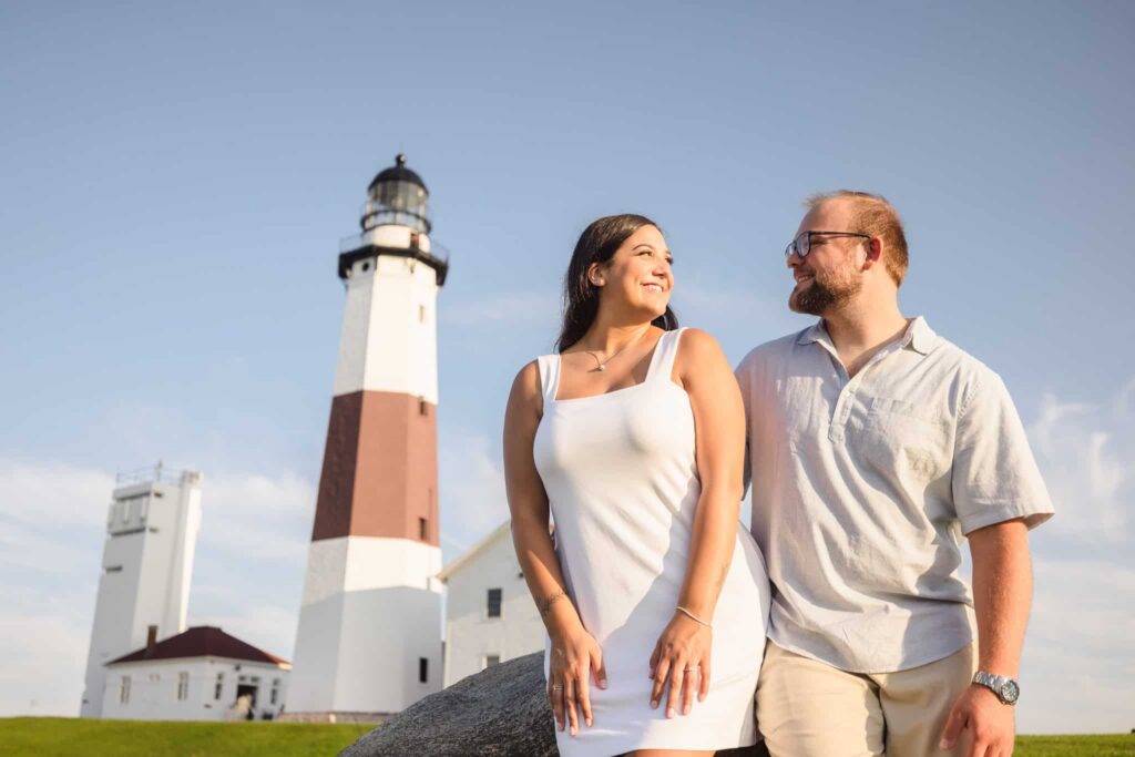 Couple seated together near Montauk Point Lighthouse during a Montauk engagement session, sharing a quiet moment with the iconic Hamptons landmark rising behind them in warm evening light.