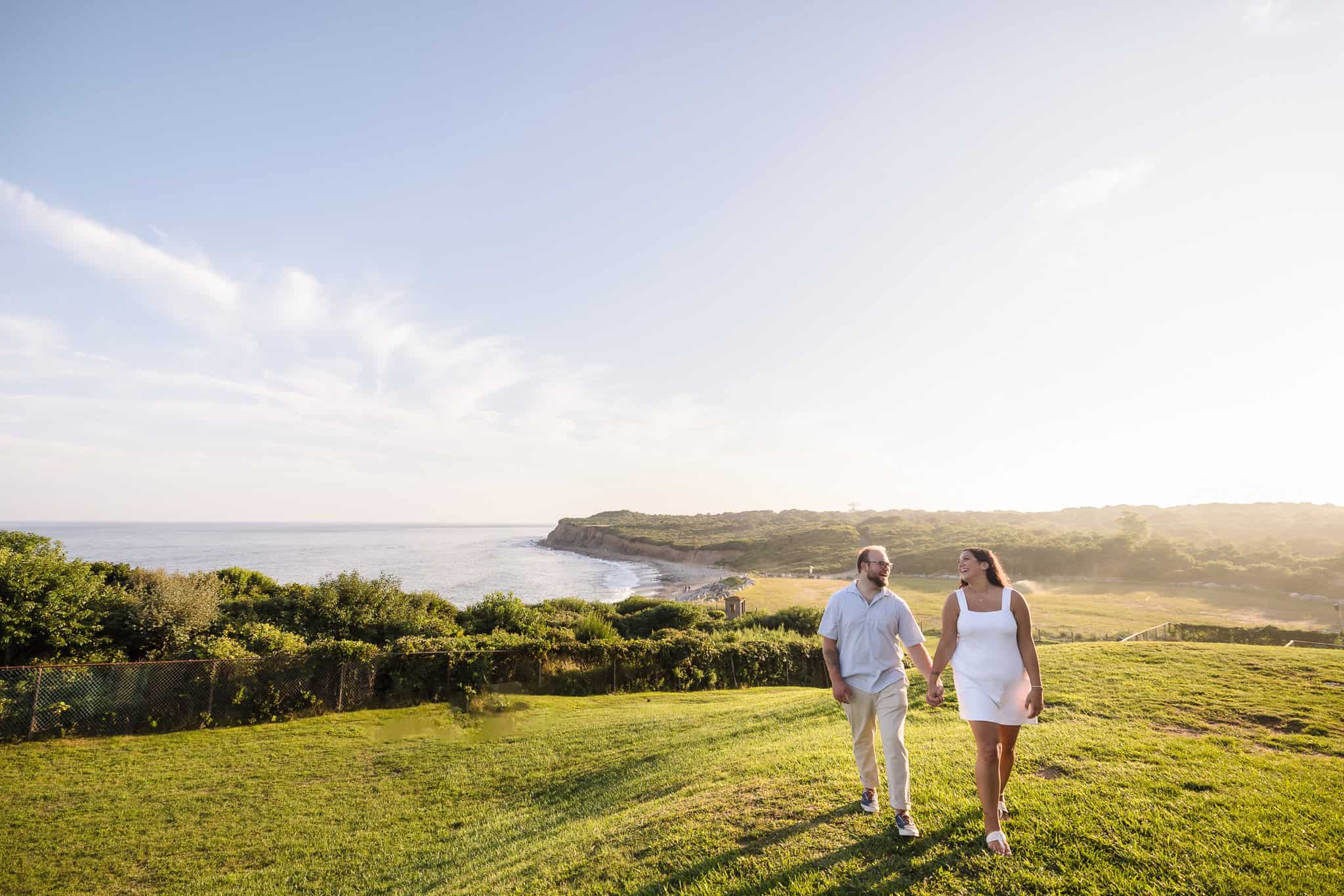 Couple walking hand in hand along the coastal bluffs during a Montauk engagement session at Camp Hero State Park, with sweeping Atlantic views and golden hour light over the East End.