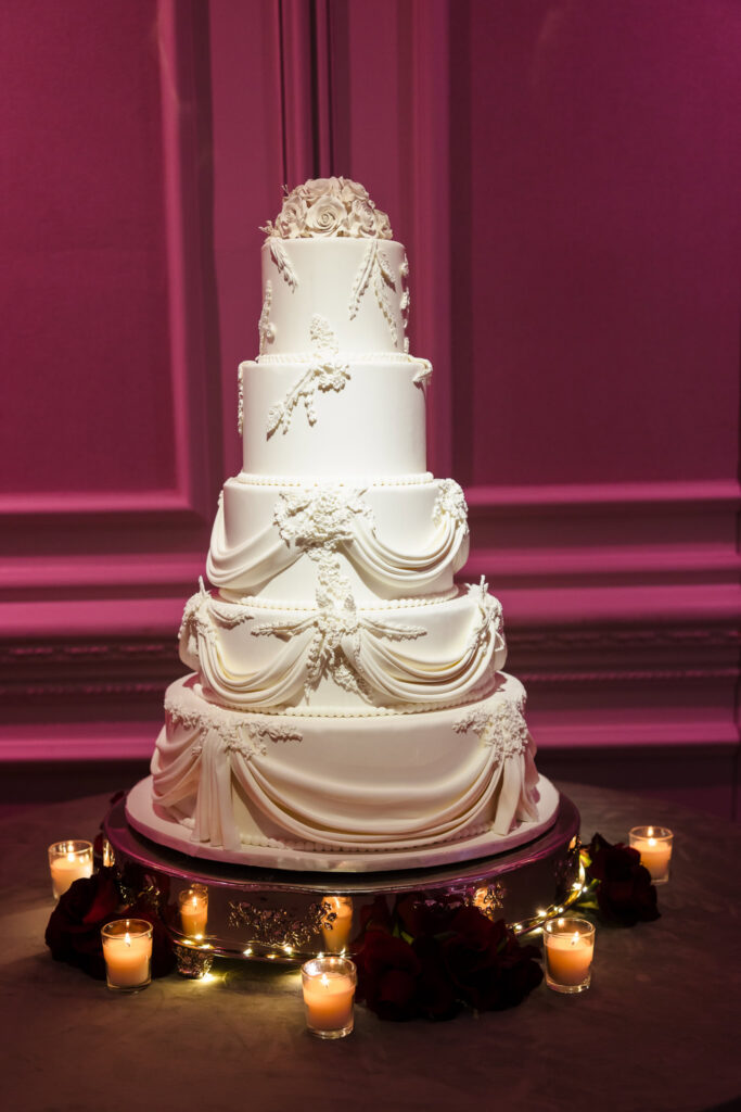 Four-tier white wedding cake with ornate draping details displayed in soft blush lighting inside thE ballroom during the Blush Hour wedding showcase.