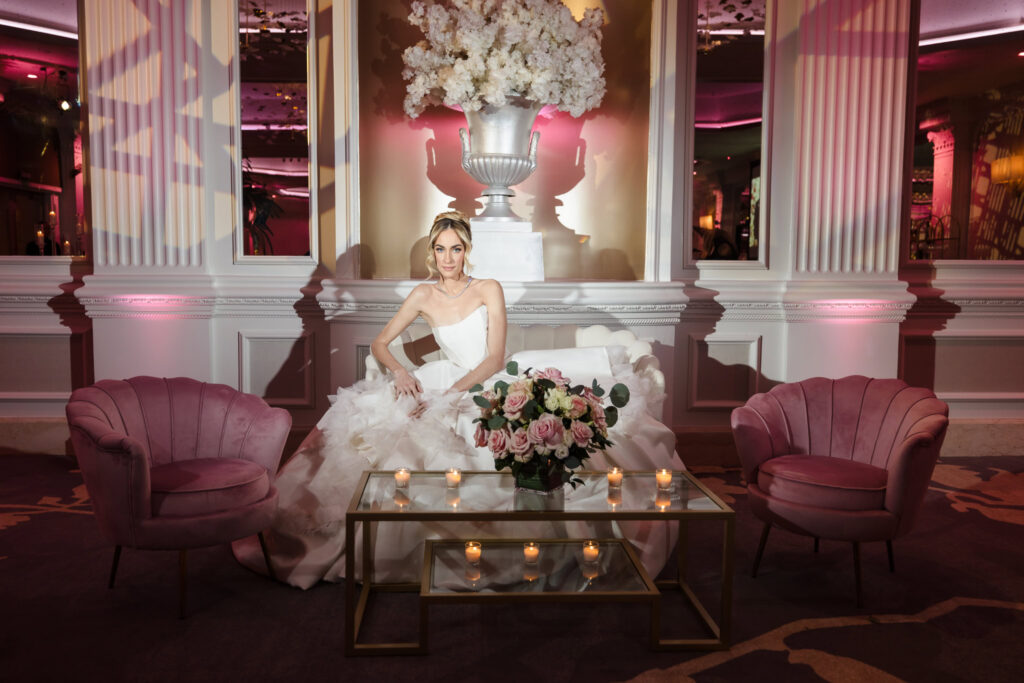 Bridal model seated in a styled lounge vignette with blush chairs, candles, and florals inside the Garden City Hotel ballroom during the Blush Hour wedding showcase.