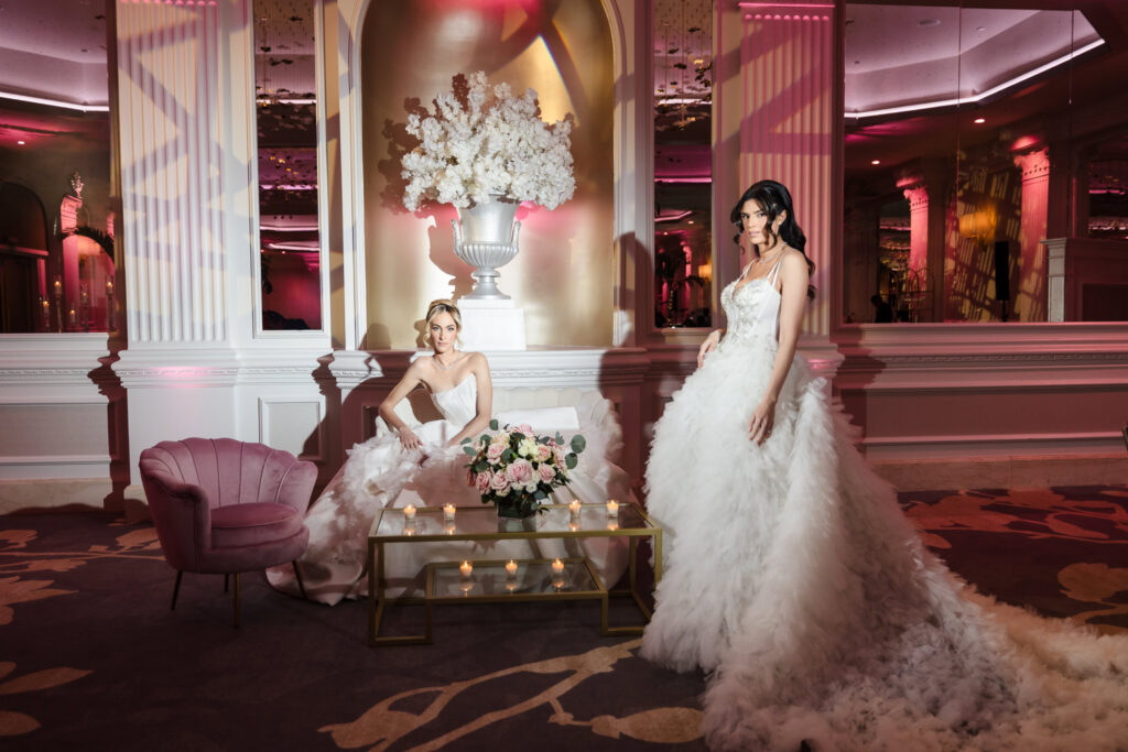Two bridal models in couture wedding gowns styled in a lounge setting inside the Garden City Hotel ballroom during the Blush Hour wedding showcase in Garden City, NY.