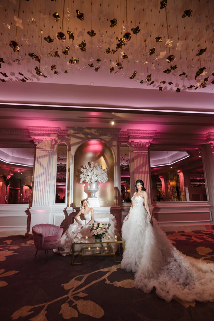 Two bridal models in elegant wedding gowns posed beneath the sculptural chandelier installation inside the Garden City Hotel ballroom during the Blush Hour event in Garden City, NY.
