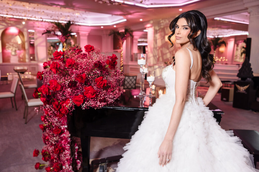 Bridal model in a textured wedding gown standing beside a grand piano with cascading red rose florals inside the Garden City Hotel ballroom during the Blush Hour wedding showcase.
