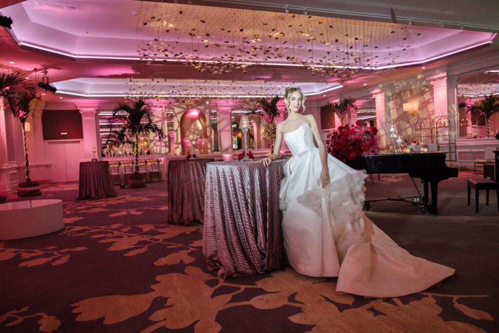 Bridal model in a strapless wedding gown leaning against a cocktail table inside the Garden City Hotel ballroom, illuminated with blush lighting during the Blush Hour wedding showcase.