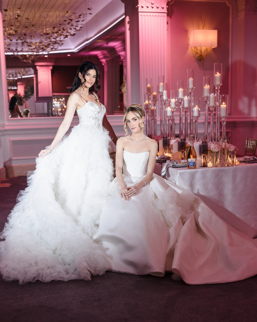 Bridal models in couture wedding gowns posing beside an elegant candlelit reception table during the Blush Hour showcase at the Garden City Hotel ballroom in Garden City, NY.