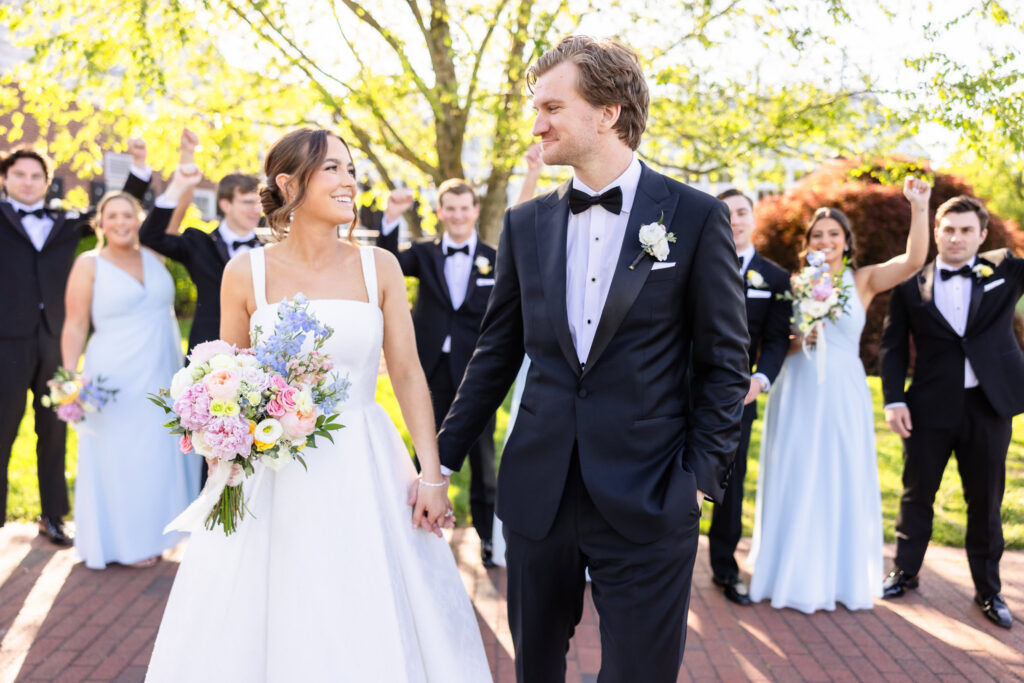 Bride and groom holding hands with their wedding party celebrating behind them during a joyful Long Island wedding.